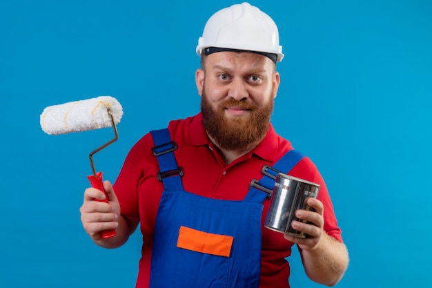 Kostenloses Foto junger bärtiger baumeistermann in der bauuniform und im sicherheitshelm, der farbroller hält und kann mit unglücklichem gesicht