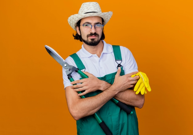 Junger bärtiger Gärtnermann, der Overall und Hut hält, die Heckenscheren und Gummihandschuhe hält, die vorne mit ernstem Gesicht über orange Wand stehen