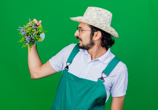 Junger bärtiger Gärtnermann, der Overall und Hut hält Topfpflanze hält, die es unzufrieden betrachtet