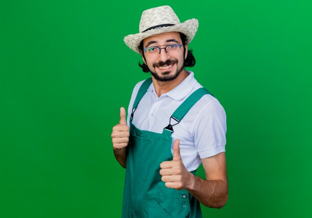 Junger bärtiger Gärtnermann, der Overall und Hut trägt, der vorne lächelnd mit glücklichem Gesicht zeigt, das Daumen oben steht über grüner Wand