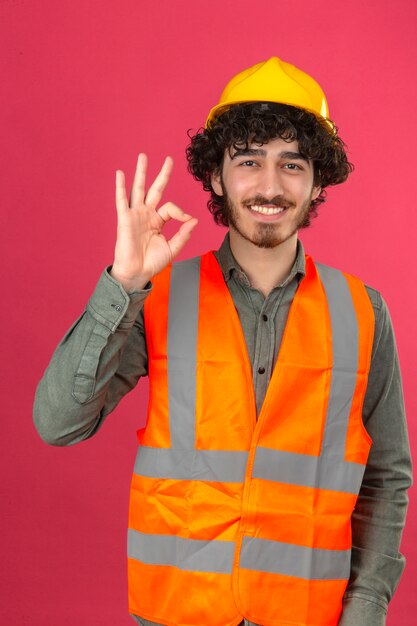 Junger bärtiger gutaussehender Ingenieur, der Sicherheitshelm und Weste trägt, die das OK-Zeichen tut, das über isolierter rosa Wand steht