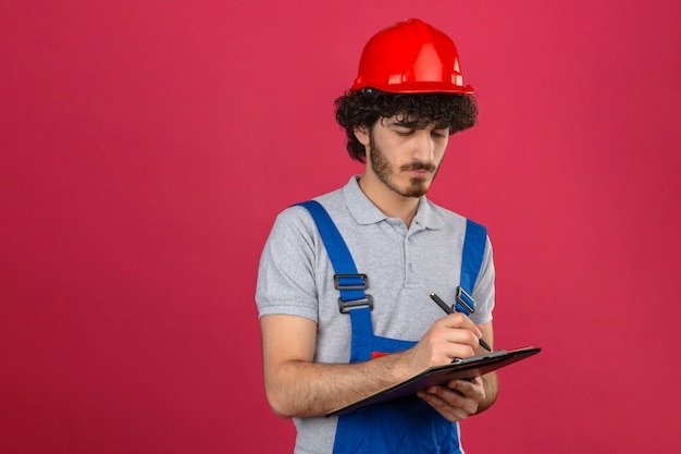 Junger bärtiger hübscher Baumeister, der Bauuniform und Sicherheitshelm hält, der Klemmbrett und Stift hält, die etwas mit ernstem Gesicht über isolierter rosa Wand schreiben