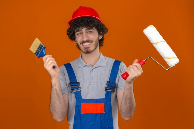 Kostenloses Foto junger bärtiger hübscher baumeister, der bauuniform und sicherheitshelm hält farbroller und pinsel in händen trägt, die glücklich über isolierte orange wand lächeln