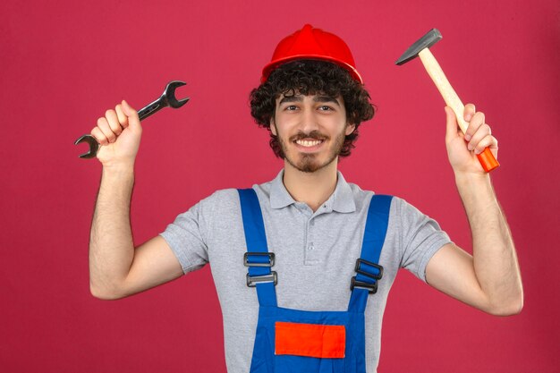 Junger bärtiger hübscher Baumeister, der die Bauuniform und den Sicherheitshelm trägt, die mit erhobenen Armen stehen, die Schraubenschlüssel und Hammer halten, die fröhlich über isolierte rosa Wand lächeln