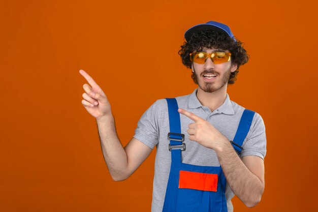 Junger bärtiger hübscher Baumeister, der eine einheitliche Brille und eine Kappe der Bauuniform trägt, die lächeln und die Kamera betrachten, die mit zwei Händen und Fingern zur Seite über isolierte orange Wand zeigt