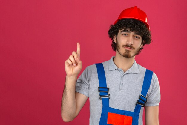 Junger bärtiger hübscher Baumeister, der Konstruktionsuniform und Sicherheitshelm trägt, der zuversichtlich zeigt Zeigefinger über isolierte rosa Wand zeigt