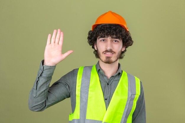 Junger bärtiger hübscher Ingenieur, der Grußgeste macht, die mit der Hand winkt, die über isolierte grüne Wand lächelt