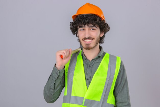 Junger bärtiger hübscher Ingenieur, der Sicherheitshelm und Weste trägt, die mit Hammer auf Schulter stehen und fröhlich über isolierte weiße Wand lächeln