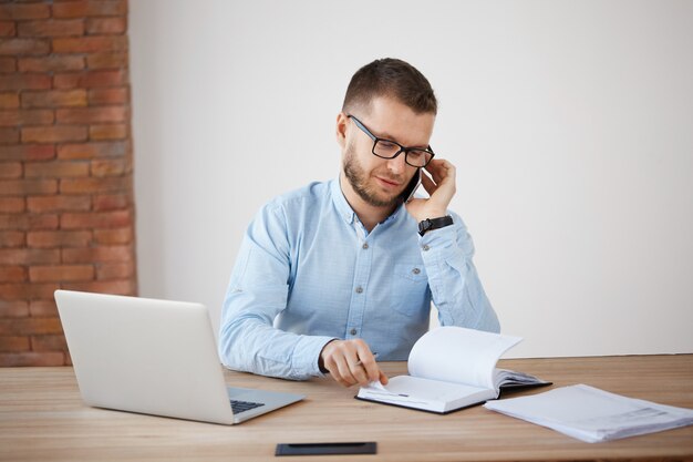 Junger bärtiger kaukasischer Manager in den Gläsern und im blauen Hemd, die mit Kunden am Telefon sprechen und Bestelldetails besprechen