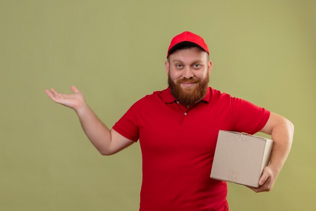 Junger bärtiger Lieferbote in der roten Uniform und in der Kappe, die den Pappkarton hält, der Kamera mit dem selbstbewussten Lächeln betrachtet, das mit Arm seiner Hand präsentiert