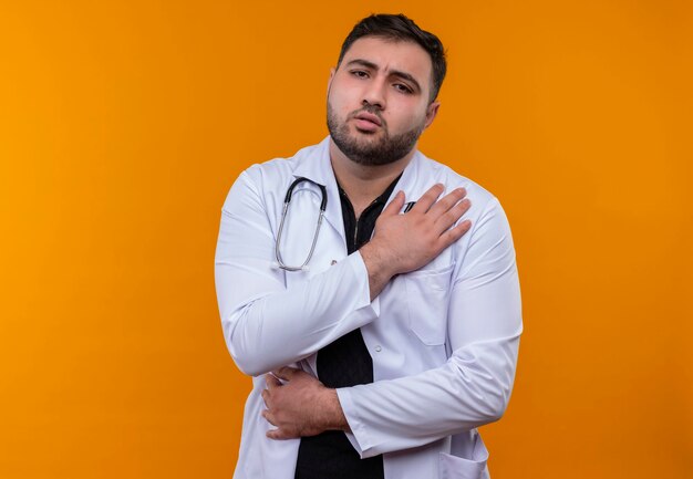 Junger bärtiger männlicher Arzt, der weißen Kittel mit berührendem Schultergefühlschmerz trägt