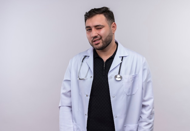 Junger bärtiger männlicher Arzt, der weißen Kittel mit dem Stethoskop zwinkert, das lächelnd Kamera betrachtet