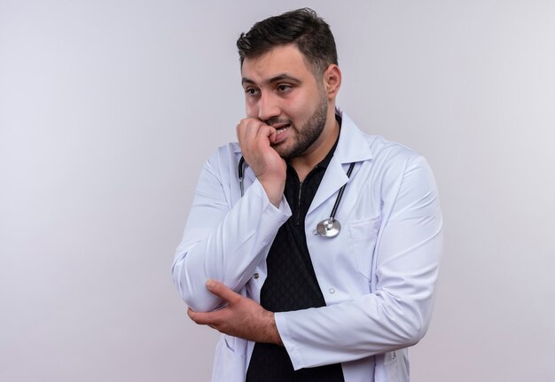 Junger bärtiger männlicher Arzt, der weißen Kittel mit gestresstem und nervösem Stethoskop seiner Nägel trägt