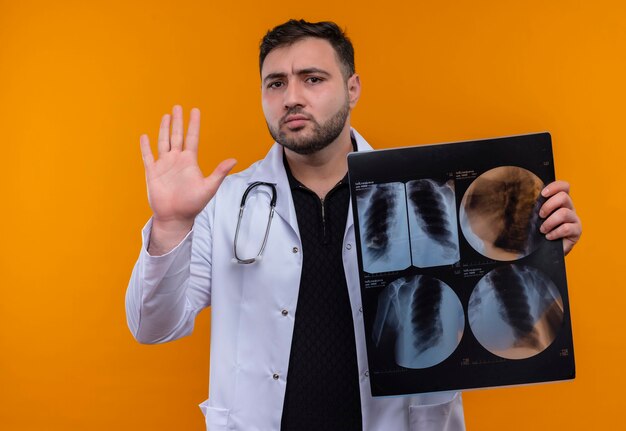 Junger bärtiger männlicher Arzt, der weißen Kittel mit Stethoskop hält, das Röntgenbild der Lunge hält, das Stoppschild mit Hand macht