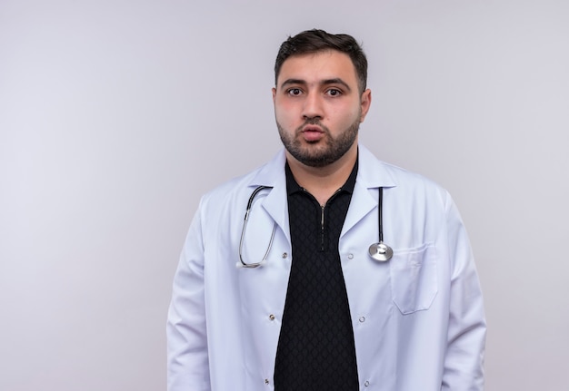 Junger bärtiger männlicher Arzt, der weißen Kittel mit Stethoskop trägt, das Kamera verwirrt betrachtet
