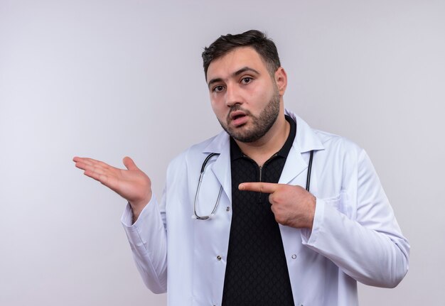 Junger bärtiger männlicher Arzt, der weißen Kittel mit Stethoskop trägt, das mit Arm oh seine Hand zeigt, die mit Zeigefinger zu Arm zeigt