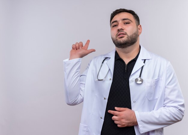 Junger bärtiger männlicher Arzt, der weißen Kittel mit Stethoskop trägt, das mit ernstem sicherem Ausdruck beiseite schaut, der auf etwas hinter zeigt