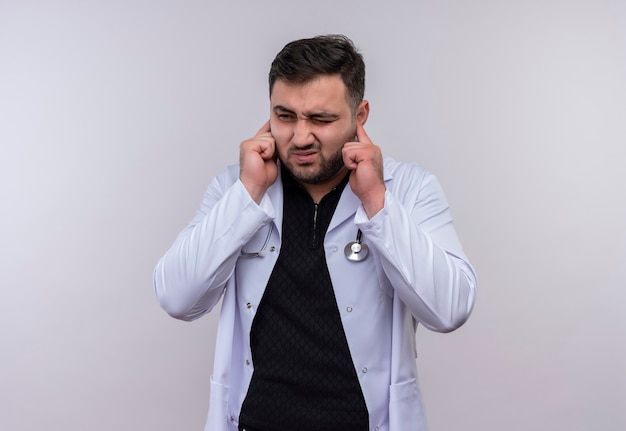 Junger bärtiger männlicher Arzt, der weißen Kittel mit Stethoskop trägt, das Ohren mit den Fingern mit genervtem Ausdruck für das Geräusch des lauten Geräusches schließt