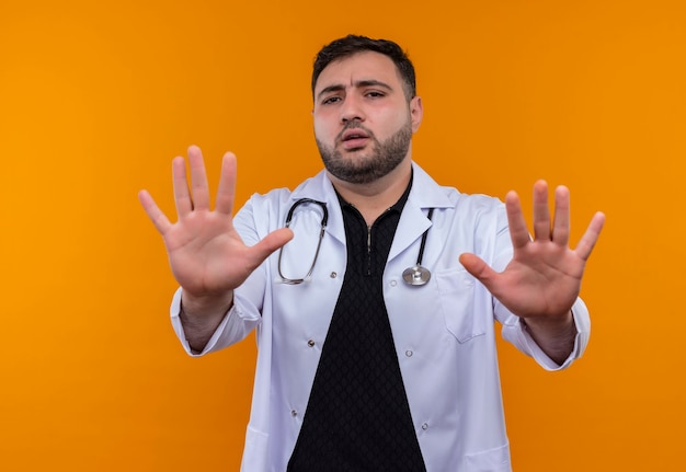Junger bärtiger männlicher Arzt, der weißen Kittel mit Stethoskop trägt, das seine Hände heraushält und Stoppschild mit Angstausdruck macht