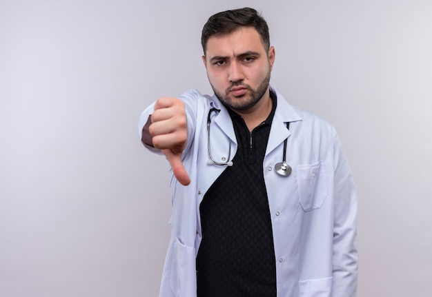 Junger bärtiger männlicher Arzt, der weißen Kittel mit Stethoskop trägt, das unzufrieden aussieht und Abneigung zeigt
