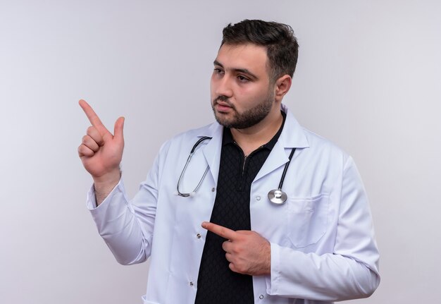 Junger bärtiger männlicher Arzt, der weißen Kittel mit Stethoskop trägt, das zur Seite schaut, mit ernstem selbstbewusstem Ausdruck, der zur Seite zeigt