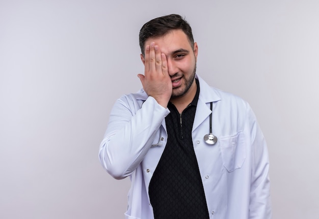 Junger bärtiger männlicher Arzt, der weißen Mantel mit lächelndem Stethoskop trägt und ein Auge mit Handfläche bedeckt
