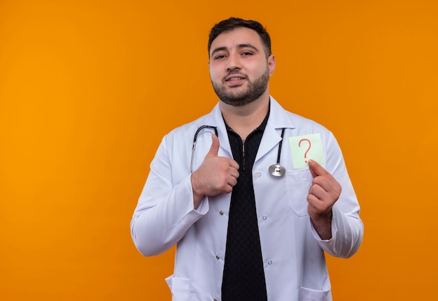 Junger bärtiger männlicher Arzt, der weißen Mantel mit Stethoskop hält, das Erinnerungspapier mit Fragezeichen lächelnd zeigt Daumen hoch