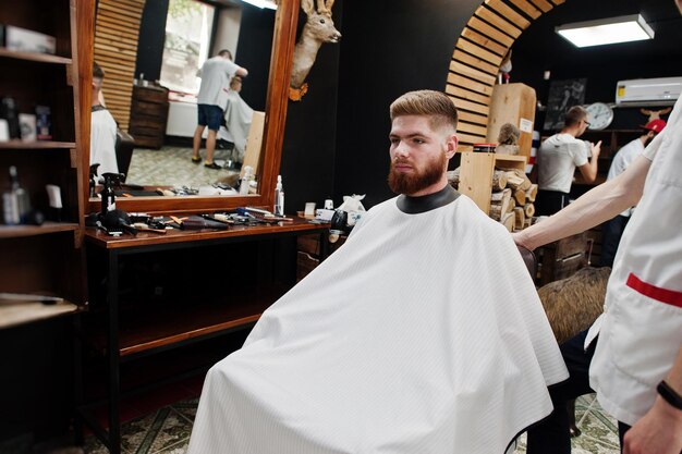 Junger bärtiger Mann, der sich vom Friseur die Haare schneiden lässt, während er im Friseursalon auf dem Stuhl sitzt Barber Soul