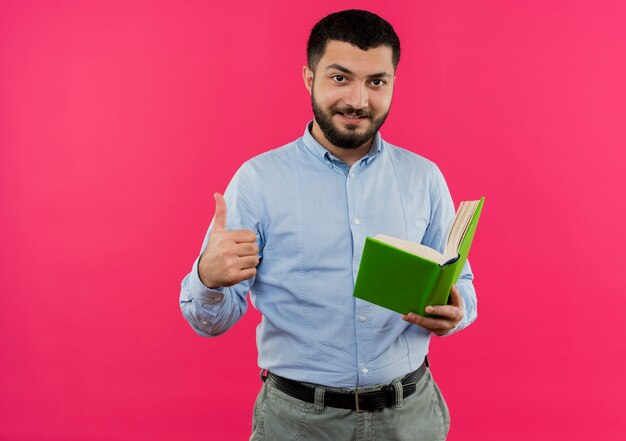 Junger bärtiger Mann im blauen Hemd, das lächelndes Buch hält Daumen zeigt