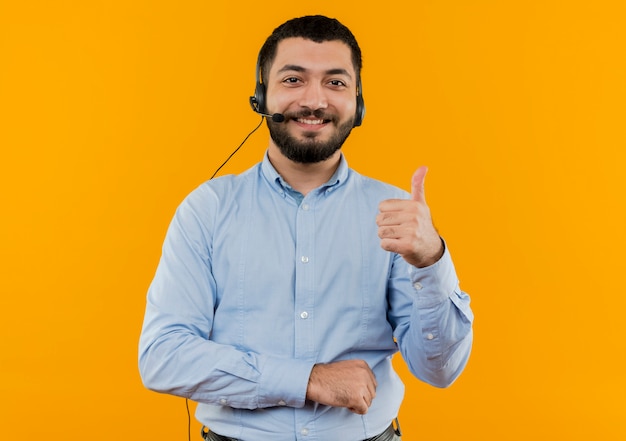 Junger bärtiger Mann im blauen Hemd mit Kopfhörern mit dem lächelnden Mikrofon, das Daumen oben zeigt