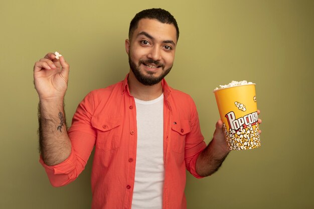 Junger bärtiger Mann im orangefarbenen Hemd, der Eimer mit Popcorn hält, der fröhlich über heller Wand stehend lächelt