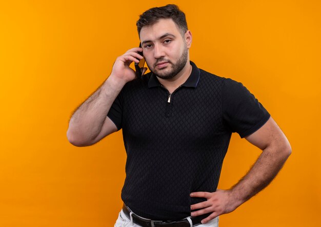 Junger bärtiger Mann im schwarzen Hemd, der beim Sprechen auf dem Handy unzufrieden aussieht