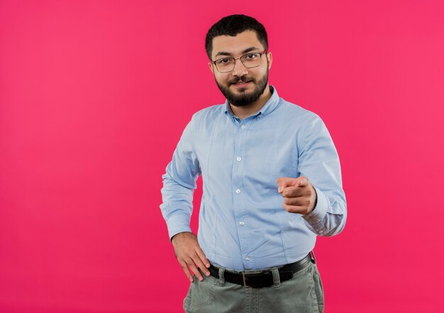 Junger bärtiger Mann in den Gläsern und im blauen Hemd, die mit Finger auf Kamera lächeln, die mit glücklichem Gesicht lächelt