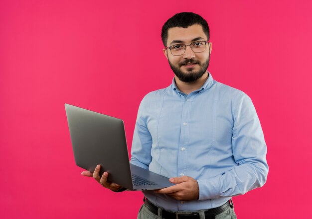 Junger bärtiger Mann in den Gläsern und im blauen Hemd hält Laptop, der zuversichtlich lächelt