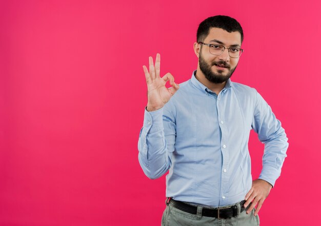 Junger bärtiger Mann in den Gläsern und im blauen Hemd lächelnd zeigt ok singen