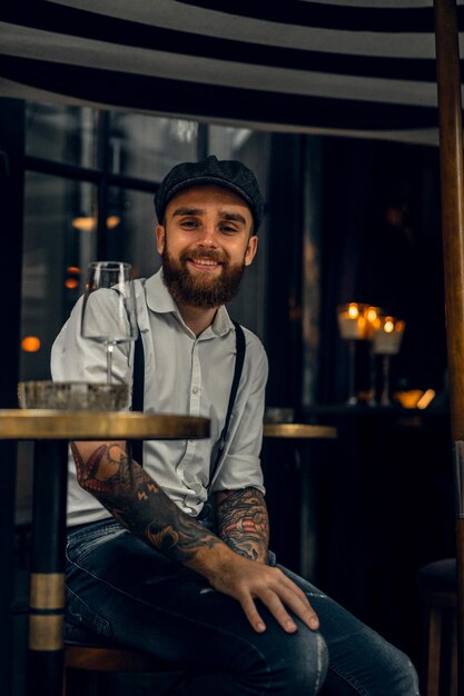 Junger bärtiger Mann in einem Café auf der Straße mit einem Glas Wein. Romantischer Typ mit weißer Hemdmütze und Hosenträgern in der Stadt. Peaky Blinders. alte fassion retro.