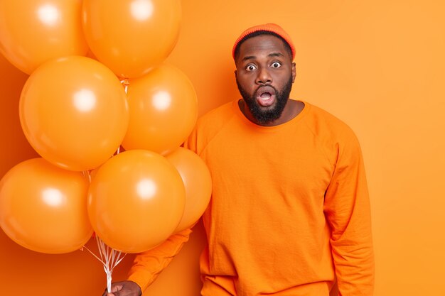 Junger bärtiger schwarzer bärtiger Mann starrt schockiert hält Mund offen vor Überraschung und Unglaube trägt orange Hut und Pullover posiert mit Heliumballons kommt auf Party isoliert drinnen