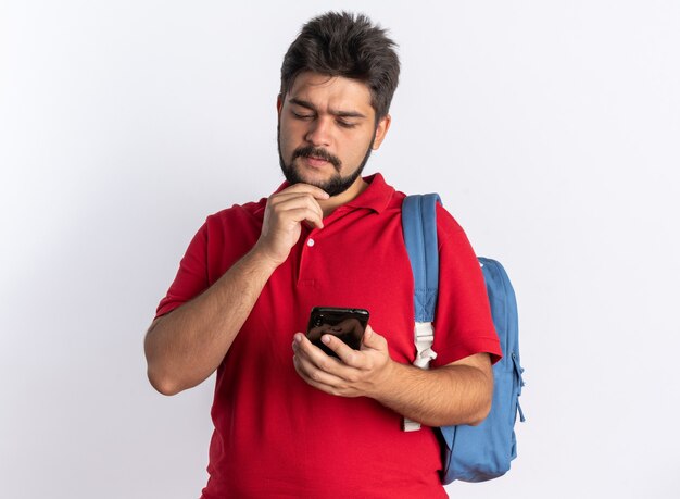 Junger bärtiger Student im roten Poloshirt mit Rucksack, der das Smartphone hält und es verwirrt im Stehen betrachtet