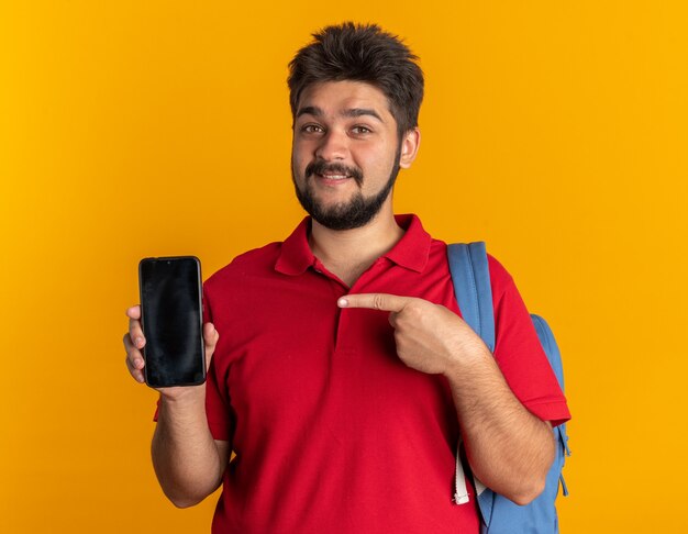 Junger bärtiger Student im roten Poloshirt mit Rucksack, der das Smartphone hält und mit dem Zeigefinger darauf zeigt, lächelt selbstbewusst glücklich und positiv über orangefarbener Wand