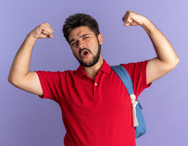 Junger bärtiger Student im roten Poloshirt mit Rucksack, der die Fäuste hebt und wie ein glücklicher und selbstbewusster Gewinner posiert