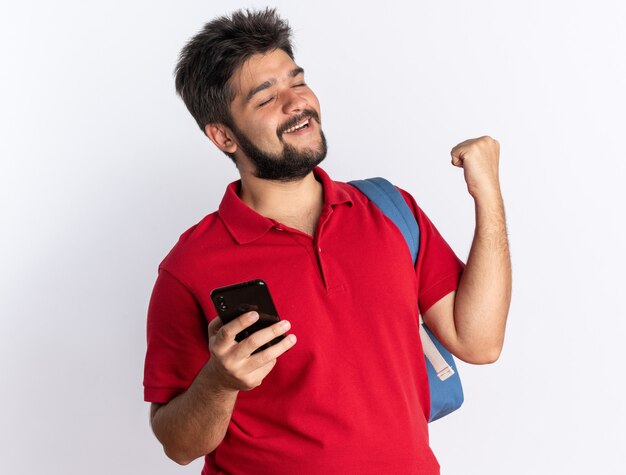 Junger bärtiger Student im roten Poloshirt mit Rucksack, der die Faust des Smartphones hält, glücklich und aufgeregt über weißer Wand stehend