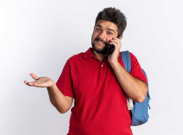 Junger bärtiger Student im roten Poloshirt mit Rucksack, der fröhlich lächelt, während er auf dem Handy über der weißen Wand spricht