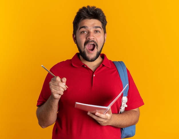 Junger bärtiger Student im roten Poloshirt mit Rucksack, der Notizbuch und Bleistift hält, erstaunt und überrascht, eine neue Idee über der orangefarbenen Wand zu haben?