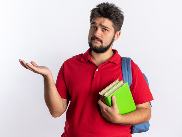 Junger bärtiger Student im roten Poloshirt mit Rucksack, der Notizbücher hält, die Kopienraum mit dem Arm der Hand präsentieren, der mit traurigem Ausdruck stehend aussieht