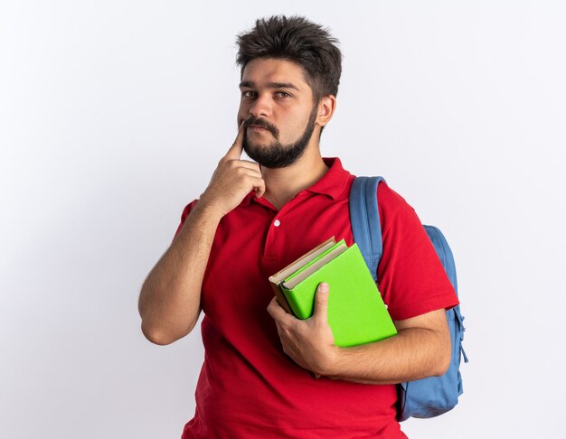 Junger bärtiger Student im roten Poloshirt mit Rucksack, der Notizbücher hält, die verwirrt über der weißen Wand stehen?