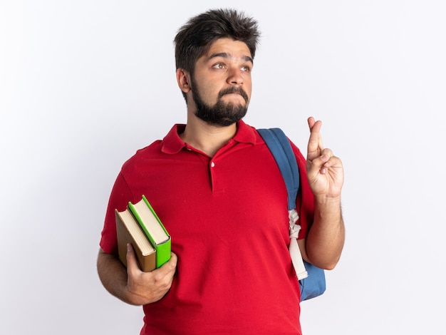 Junger bärtiger Student im roten Poloshirt mit Rucksack, der Notizbücher hält, die Wünsche mit Hoffnungsausdruck machen, der die Finger stehend kreuzt