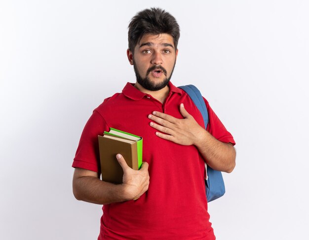 Junger bärtiger Student im roten Poloshirt mit Rucksack, der Notizbücher hält, überrascht, die Hand auf seiner Brust zu halten, die über weißer Wand steht?