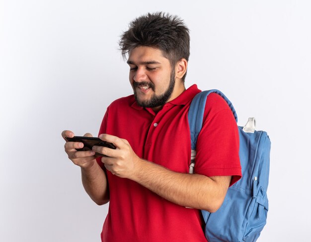 Junger bärtiger Student im roten Poloshirt mit Rucksack, der Spiele mit Smartphone spielt, glücklich und fröhlich stehend