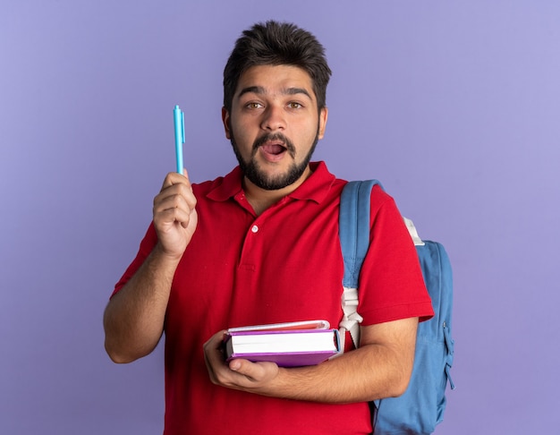 Junger bärtiger Student in rotem Polohemd mit Rucksack, der Bücher und Stift hält, überrascht mit einer neuen Idee, die über blauer Wand steht?