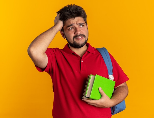 Junger bärtiger Student in rotem Polohemd mit Rucksack, der Notizbücher hält, die beiseite schauen, verwirrt mit der Hand auf dem Kopf für Fehler im Stehen